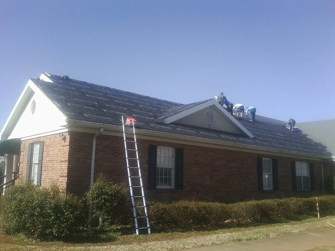 Crew tearing shingles off a brick ranch roof with a ladder set — Metal Roof Repair work by our crew in Evans City