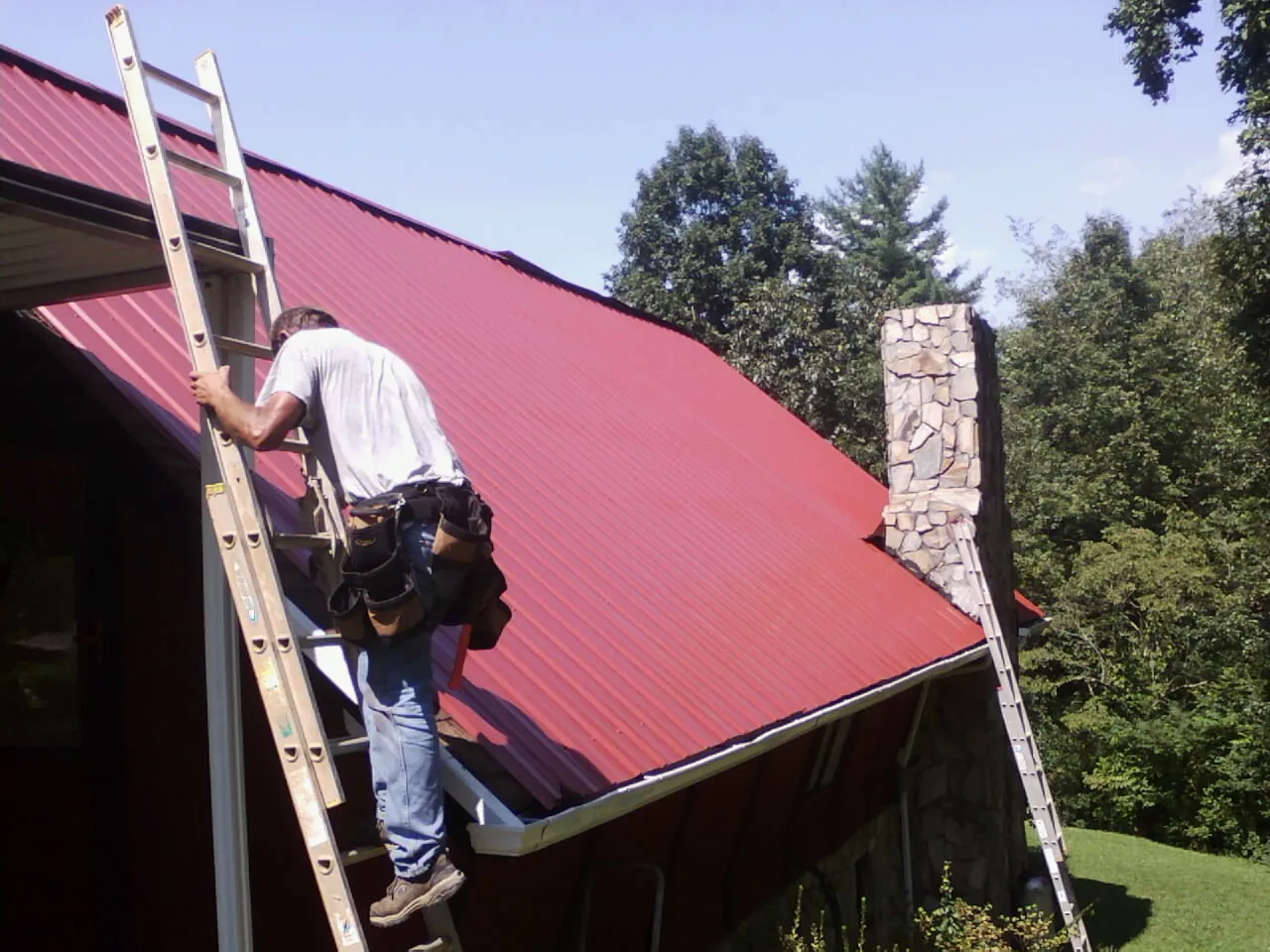 Roofer on a ladder beside a red metal roof and stone chimney — Soffit & Fascia Repair by Roofing Brothers in Evans City